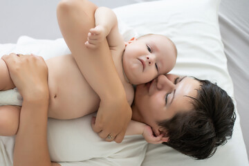 Mother and child on a white bed. Mom and baby boy in diaper playing in bedroom. Parent and little kid relaxing at home. Family having fun together.