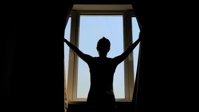 Back view: silhouette of woman opening curtains, looking out of window and stretching her arms in room at morning