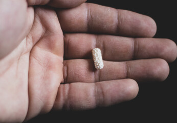 transparent pill with small white spheres inside, held by left hand of brown-skinned male, on black background