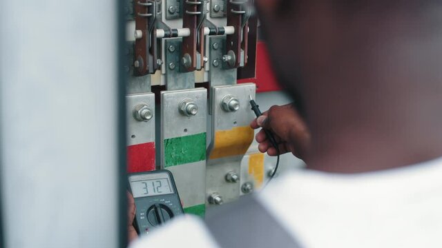 Close up of african american man standing near switchgear compartment with multimeter in hands. Competent technician controlling working process of solar station
