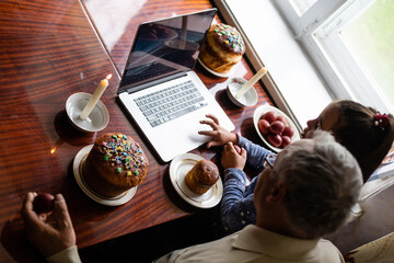 grandfather and granddaughter are talking via video link to their friends. Decorated table with colorful eggs and cake. Chatting during the COVID pandemic and the Easter holidays