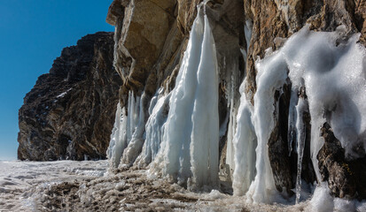 Fototapeta na wymiar Early spring. Huge icicles at the base of the rocks begin to melt. Loose melted snow. A sunny day. Clear blue sky. Baikal