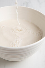 Nut milk bag squeezing milk into a white bowl with almond milk placed on a white marble surface
