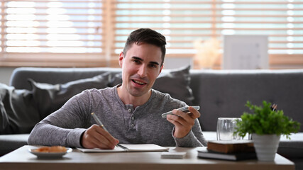Smiling man making notes on notebook and talking on mobile at home.