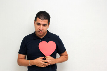 Latino adult man with a big red heart shows his anger, disgust, sadness for the arrival of February and celebrating Valentine's Day of Love and Friendship with his family, partner and friends
