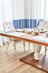 Luxury vintage dining table of walnut solid and veneer wood with festive setting and armchairs near window in spacious room close-up