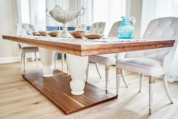 Luxury vintage dining table of walnut solid and veneer wood with festive setting and armchairs near window in spacious room close-up