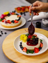 Pouring blueberry sauce onto delicious sweet cheesecake decorated with fresh berry fruits, and flowers on a wooden plate, for a birthday, Valentine day and celebration