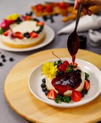 Pouring blueberry sauce onto delicious sweet cheesecake decorated with fresh berry fruits, and flowers on a wooden plate, for a birthday, Valentine day and celebration
