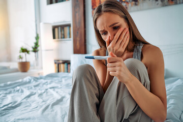 Excited young girl in home clothes sitting on the bed and looking sadly at the pregnancy test covering her mouth with her hand in surprise.