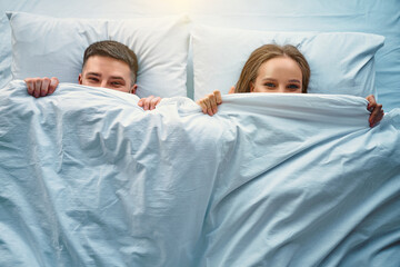 The cheerful couple is lying in bed and covered with a blanket half face and laughing. Playful emotions of joy, cunning and cheerfulness. Top view.