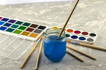 watercolor paints a jar of water and brushes for drawing on a gray textured surface