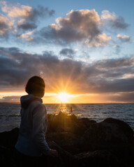 Woman looking at the sunrise on the rock beach with sun shining through the clouds. Vertical format.