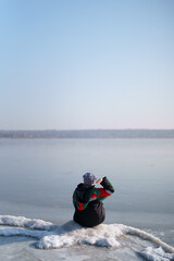 Fototapeta na wymiar A woman in a colorful thermal costume enjoying winter while sitting and watching a frozen lake with blue sky in the background. Freedom