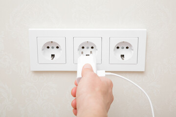 Young adult woman hand holding and plugging white electrical plug for smartphone charging in wall...