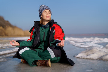Fototapeta na wymiar A woman in a colorful thermal costume enjoying winter while sitting in a meditation pose on a frozen lake with blue sky in the background. Freedom, peace, quiet, serenity
