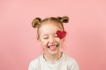 Funny little blonde girl smiling and playing with red heart toy on pink background. Valentines Day or Mothers Day concept