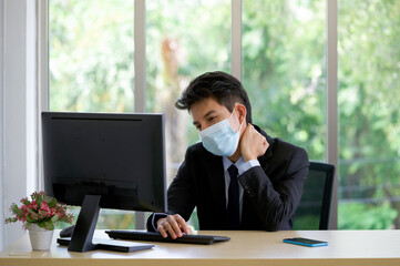 Young asian businessman in suit and face mask grab the neck with his hand. To alleviate pain from...