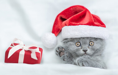 Cute kitten wearing red santa's hat lying with gift box under a white blanket on a bed. Top down view