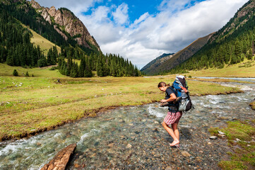 Fototapeta premium Young woman with big backpack barefoot crossing river. Shoeless female backapcker in river. Karakol valley, Issyk-kul region, Ala-kul lake Terskey Alatau mountain range, Kyrgyzstan, Central Asia.