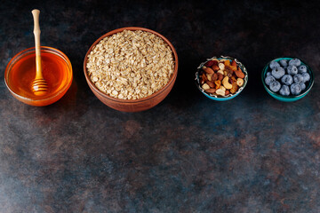 Bowl with oatmeal flakes, nuts, berries and honey on dark background. Ingredients for healthy breakfast with oatmeal and blueberries. Copy space. Top view