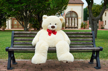 big polar plush bear with red tie, sits on a bench - obrazy, fototapety, plakaty