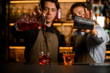 two clear glasses with ice in which bartenders carefully pour different cocktails