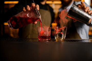 Close-up view of two transparent glasses with ice, into which bartenders carefully pour various steaming cocktails