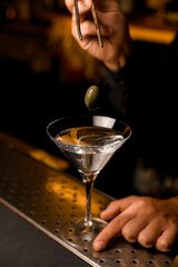 focus on green berry olive over transparent martini glass with cocktail on bar counter