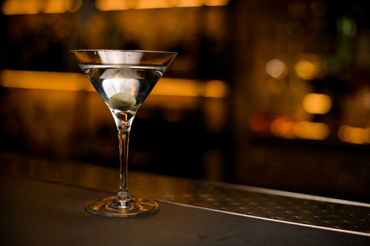 martini glass with beverage and green olive on bar counter. Blurred bar background.