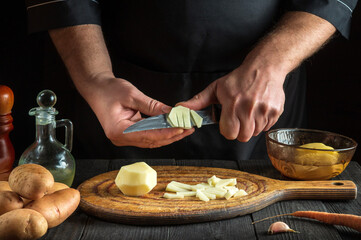 The chef uses a knife to cut the raw potatoes into small pieces. Close-up of a cook hands while working in a restaurant kitchen.