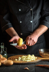 Chef cuts raw potatoes into pieces with a knife before preparing breakfast or dinner. Close-up of cook hands while working in a restaurant kitchen