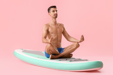 Young man meditating on sup board against color background