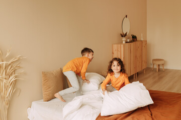 Happy cheerful sleepy children brother and sister in orange bright pajamas have fun laughing and fighting with pillows in a cozy bedroom at home. Children's pajama party. Selective focus