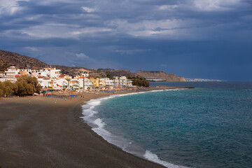 Fototapeta premium Wide sandy beach and Mediterranean town by the sea under dramatic clouds, Myrtos, Crete, Greece