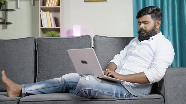 Wide angle shot of relaxed young man busy using laptop while sitting on safa at home - concept of work from home, new normal employee or job lifestyle