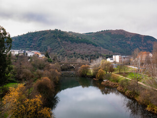 Fototapeta na wymiar View of the Sil river as it passes through the city of Ponferrada with vegetation around it and the train tracks on a bridge and the Pajariel in the background on a cloudy day