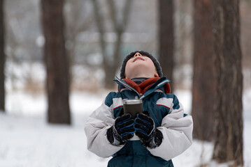 Portrait of child with mug in his hands on winter forest background. Boy looks up at the sky.