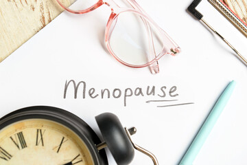 Clipboard with word MENOPAUSE, eyeglasses and alarm clock on light wooden background, closeup