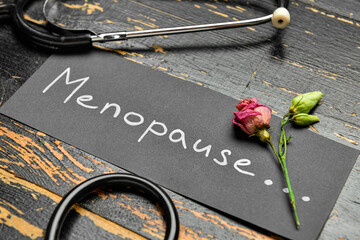 Paper with word MENOPAUSE, dried rose and stethoscope on dark wooden background, closeup