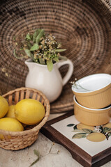 Yellow lemons in a straw basket near the stand under hot with painted lemons, ceramic molds and a ceramic jug with dry lime blossoms against a background of a rattan tray