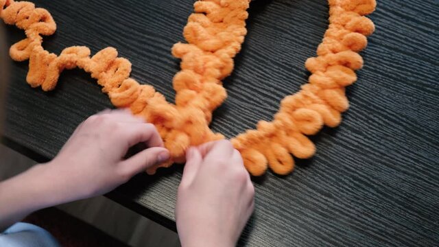 The girl knits with her hands a balaclava of orange color with the help of Alize Puffy threads. Top view.