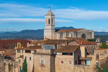 Fototapeta na wymiar Girona Cathedral rises above the tiled roofs of the Gothic Quarter in the Old Town. Medieval cityscape with ancient buildings against the backdrop of mountains and blue sky on a sunny autumn day
