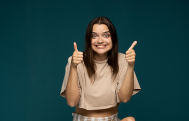 Happy young brunette female wearing beige oversize t-shirt making thumb up sign and smiling cheerfully, showing her support and respect to someone. Body language. I like that. Good job.