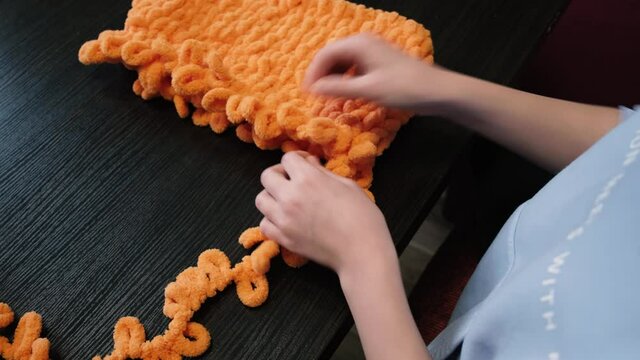 The girl knits with her hands a balaclava of orange color with the help of Alize Puffy threads. Half the work is done.