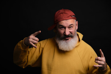 Studio half-length portrait mature bearded man in yellow hoodie and baseball cap shows rock'n'roll...