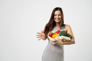 Happy smiling woman in grey dress holding reusable net bag with fruits and vegetables on white background. Food shopping and eco friendly concept.