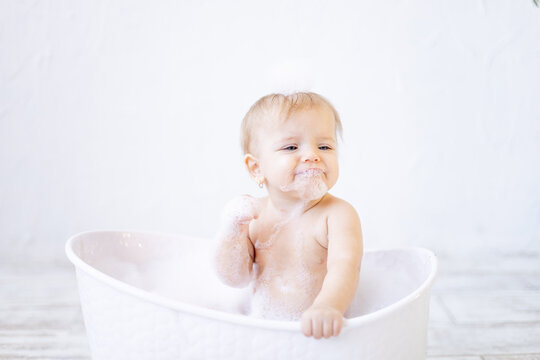 a crying little baby girl with foam in her mouth and in front of her eyes bathes in a bathtub with foam and soap bubbles in a bright bathroom, the concept of child hygiene