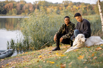 Pleased asian and latin man sitting, caressing his Maremmano-Abruzzese Sheepdog and looking away in nature at sunny autumn day. Concept of rest and weekend in nature