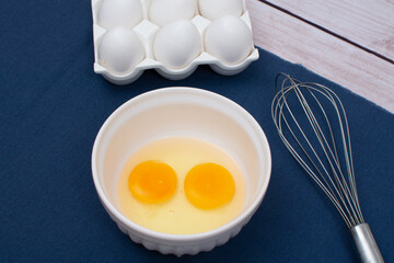 Egg preparation for recipe, with broken eggs, egg tray and a fouet on a blue towel and a wooden table, top view